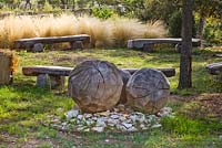 Un cercle de bancs en bois de Marc Nucera en forêt avec Stipa tenuissima et sculpture en boule en bois également de Marc Nucera. Provence, France, Domaine de la Verriere