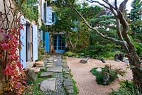 Jardin de style japonais - vue sur le côté est de la maison avec volets bleus, chemin avec galets et rochers, jardin de gravier et pin sylvestre taillé
