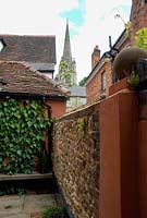 Petit jardin de ville avec un mur recouvert de lierre, un banc en bois et une bille sous un mur de silex et de briques. Pavage en pierre de York et une flèche d'église en arrière-plan
