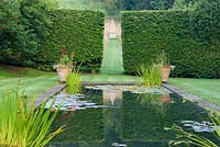 Étang formel bordé de Crataegus x lavallei standard dans le jardin supérieur où un espace entre des haies de hêtres encadre un obélisque situé dans une prairie herbeuse. Corscombe House, Corscombe, Dorset, Royaume-Uni