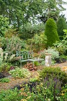 Jardin à la fin de l'été avec banc en fer forgé peint en bleu, vieux cadran solaire, chemins de gravier, roses, plantes vivaces herbeuses et vue sur l'ifiaire.
