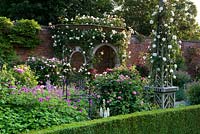Obélisque en bois et siège couvert de tonnelle en bois dans un jardin clos. Parterres de fleurs aux bords plantés de roses et de psilostemon géranium. Seend, Wiltshire