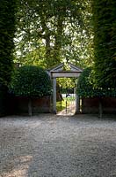 Vue depuis l'allée de style français à la porte formelle dans le jardin clos avec fronton en pierre ornée et arbres topiaires symétriques jumeaux - Seend, Wiltshire