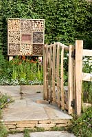 Présentez le jardin visible. La porte de canne fendue rustique s'ouvre sur un sol en pierre avec un hôtel à insectes en bois contre une haie de charme. Jardin animalier