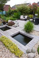 Terrasse en bois dur contemporain Ipe sur étang, texture contrastée de galets arrondis ramollis par la plantation de Miscanthus sinensis, Pittosporum ten. 'Tom Thumb '. Euonymus fortunei' Emerald 'n' Gold 'et faible couverture Buxus sempervirens