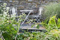 Table et chaises de café en métal contre un mur de pierre, encadrées de miscanthus et d'autres arbustes.