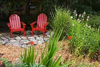 Deux chaises Adirondack en bois rouge sur un patio en dalles à côté de Miscanthus sinensis 'Strictus' - Herbe zébrée, floraison rose Physotegia virginiana - Plante obéissante, Crocosmia montbretia rouge 'Lucifer'