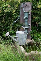 Pompe à eau en fonte montée sur socle en chêne dans une haie d'aubépine au-dessus d'une auge en pierre avec arrosoir antique.