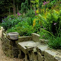 Des murs de soutènement en pierre de York se dressent entre un chemin de gravier et des parterres de fleurs en pente de digitales, fougères, centaurées, euphorbes, azalées et genêts.