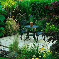Dans un petit jardin de banlieue, une cour secrète avec table et chaises, cachée derrière des cercis, des tulipiers, du phormium, des herbes et des haies de lauriers.