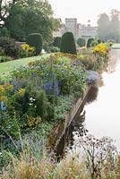 Le Long Pond avec maison au-delà, bordé par le parterre de fleurs herbacées planté d'héléniums, kniphofias, dahlias, asters et eupatoriums. Forde Abbey, nr Chard, Dorset, Royaume-Uni