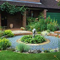 Le jardin avant a une cour en contrebas avec une sculpture centrale en bois située à heuchera. Derrière, un parterre d'iris, de phormium, de phlome, de fougère d'asperge et de giroflée.
