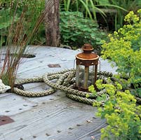 Sur une bobine de câble en bois argenté et secouru se trouve une lanterne rouillée dans une bobine de corde, par Alchemilla mollis mousseux.