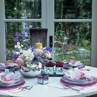 Joli vase de petits pois, de roses, de delphinium et de gypsophile au centre d'une table joliment dressée dans une véranda. Vues du jardin au-delà.