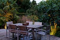 Une terrasse en bois surélevé avec mobilier de jardin pour les repas en plein air. Des Uplighters illuminent les plantations environnantes, notamment le palmier Trachycarpus fortunei, Deschampsia cespitosa 'Bronzeschlier'