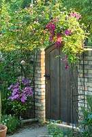 Porte de jardin en chêne encadrée par une arche recouverte de clématites et de roses. Mai