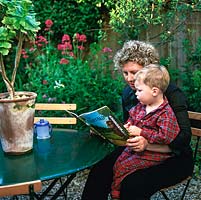 Mhairi Clutson lisant une histoire de parterre de fleurs à son plus jeune enfant, Phelin, dans leur petit jardin à l'arrière de style méditerranéen avec son patio en gravier en contrebas au-dessous d'une cour fermée.