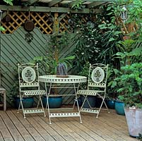 Chaise et tables sur terrasse en bois ombragée, cachées derrière des panneaux de clôture, des pots et des jasmoïdes Trachelospermum, nichées au fond d'un jardin de ville de 27 mx 13 m.
