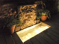 La nuit, une fontaine murale de masque de lion illuminé jette de l'eau dans la piscine en dessous, coulée dans une terrasse en bois.