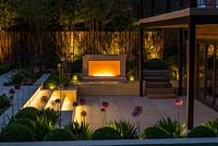 Jardin de ville conçu par Kate Gould, éclairé la nuit. Les flammes d'une cheminée à gaz ouverte illuminent la terrasse engloutie qui est bordée de parterres de boules de boîtes entrecoupées d'allium violet et blanc. La limite arrière est plantée de grands bambous dorés.