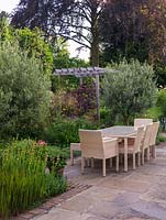 Table à manger et chaises sur terrasse ensoleillée. Parterres d'oliviers, sauge, géranium Ann Folkard, Sisyrinchium striatum, cercis, euphorbia, graminées ornementales.
