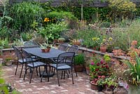 Une table et des chaises en plein air sur la terrasse en brique, avec mur de soutènement, vivaces mixtes et plantation en pot.