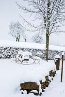 Scène d'hiver d'un jardin rural avec un mur en pierre sèche couverte de neige, des meubles de jardin, une haie et des arbres fruitiers en arrière-plan