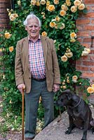 David Austin Roses. Portrait de M. David Austin OBE VMH, accompagné de son bull-terrier du Staffordshire, Bertie. Derrière, grimpant sur le mur, Rosa 'Crown Princess Margareta '.