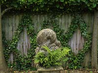 Dans un coin ombragé, sculpture en pierre sur socle planté de fougères. Derrière, une clôture terne est acclamée par un treillis de lierre.