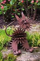 Gros plan d'une sculpture de chat en métal rouillé et de fleurs rouges d'Euphorbia milii 'Couronne d'épines' en arrière-plan dans un jardin Zen privé en arrière-cour en été