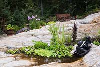 Sculpture de jeune garçon et fille en barque dans un étang dans la cavité rocheuse avec Eichornia crassipes - Jacinthe d'eau, Typha latifolia - Plantes de quenouilles communes, fleurs Phlox violettes