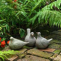 Trois sculptures d'oiseaux en céramique sont cachées sur un patio en briques sous des fougères.