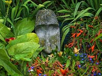 La tête en pierre recouverte de lichen se niche parmi le feuillage et les fleurs.