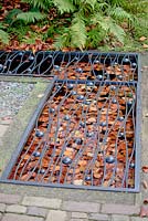 Petit étang carré rempli de feuilles d'automne brunes flottantes recouvertes d'une grille métallique décorée de noir dans le trottoir.