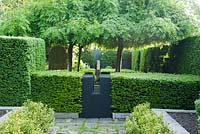 Vue sur des haies d'ifs dans une cour avec quatre cendres pleureuses dressées, Fraxinus excelsior 'Pendula' et une sculpture de David England comme point focal.