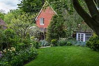 La vue sous le hêtre de Quarry Cottage à la fin du printemps. La pelouse est bordée de parterres de fleurs, dont des tulipes, des myosotis, des alliums et des balles en topiaire. Le jardin descend sur la colline jusqu'au cottage victorien.