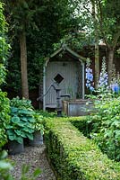 Un coin ombragé d'un jardin clos avec un siège en bois couvert, un petit étang fait d'un abreuvoir en métal galvanisé et d'une plantation tolérante à l'ombre, y compris Hosta et des haies de buis.