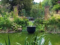 Jardin d'eau avec fontaine, jardin fleuri, pergola et pots. Piscine bordée d'alchémille, de sureau panaché, de rodgersia, d'acer, de Stipa gigantea, d'agapanthe.