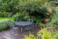 Un patio circulaire en pierre avec table et chaises. La plantation dans le parterre de fleurs surélevé environnant comprend Sedum, Achillea, Stipa gigantea, Viburnum et un arbre Sorbus.