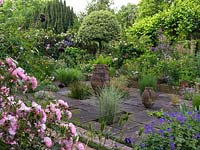 Voir devant Rosa 'Bonica' et Geranium ibericum au jardin en contrebas pavé avec des pots et des herbes.