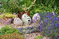 Têtes décoratives en argile dans un jardin de gravier avec Lavandula 'Hidcote' et Fuchsia 'Chequerboard', Helichrysum petiolare 'Limelight' en pots