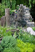 Ruine d'église aux murs de pierre, chemin, auge et plantes de jardin, y compris Digitalis purpurea - digitale et herbes. The Evaders Garden par Chorley Council. RHS Chelsea Flower Show, 2015.