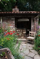 The Old Forge Artisan Garden for Motor Neurone Disease Association - RHS Chelsea Flower Show 2015. Vue des sentiers de roche naturelle et un hangar en brique et une chaise vintage entourée de fleurs sauvages
