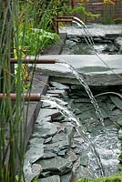 Le grand jardin Chelsea Challange Garden. Plan d'eau avec murs en ardoise, pavage, fontaines à canal et à canalisation. RHS Chelsea Flower Show, 2015