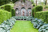 Pelouse bordée d'hostas et vue sur le mur avec des niches et trois personnages. Jardin Plas Brondanw, Pays de Galles. Conçu par et était la maison de Sir Clough Williams-Ellis. Juillet 2015