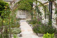 Parterres de plantes herbacées bordées de bois avec des obélisques en acier rouillé supportant des roses et des clématites.