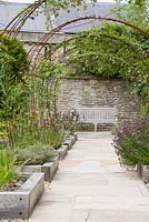 Parterres de plantes herbacées bordées de bois avec des obélisques en acier rouillé supportant des roses et des clématites.