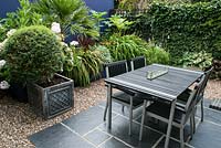Une salle de jardin conçue pour recevoir avec un pavé gris anthracite surmonté d'une table et de chaises, un mur de cobalt, une clôture recouverte de lierre et des feuilles audacieuses de palmiers et de cordylines.
