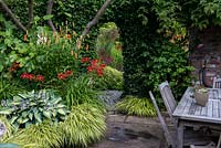 Un mur de briques recouvert de lierre avec porte mène à une zone herbacée. Au premier plan, le parterre est planté d'hostas, de graminées Hakonechloa, d'hémérocalles rouges et de Lilium martagon orange.