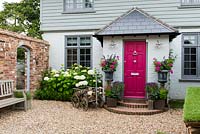 Cottage avec porte d'entrée flanquée d'une paire d'urnes plantées de Fuchsia 'Sunray', Salvia 'Black Knight', Pelargonium hortorum 'Orbit Synchro' et cosmos rose. Parterre de fleurs gauche planté d'hortensia 'Annabelle' et haie de buis. La porte dans le mur de briques à gauche mène au petit jardin de la cour.